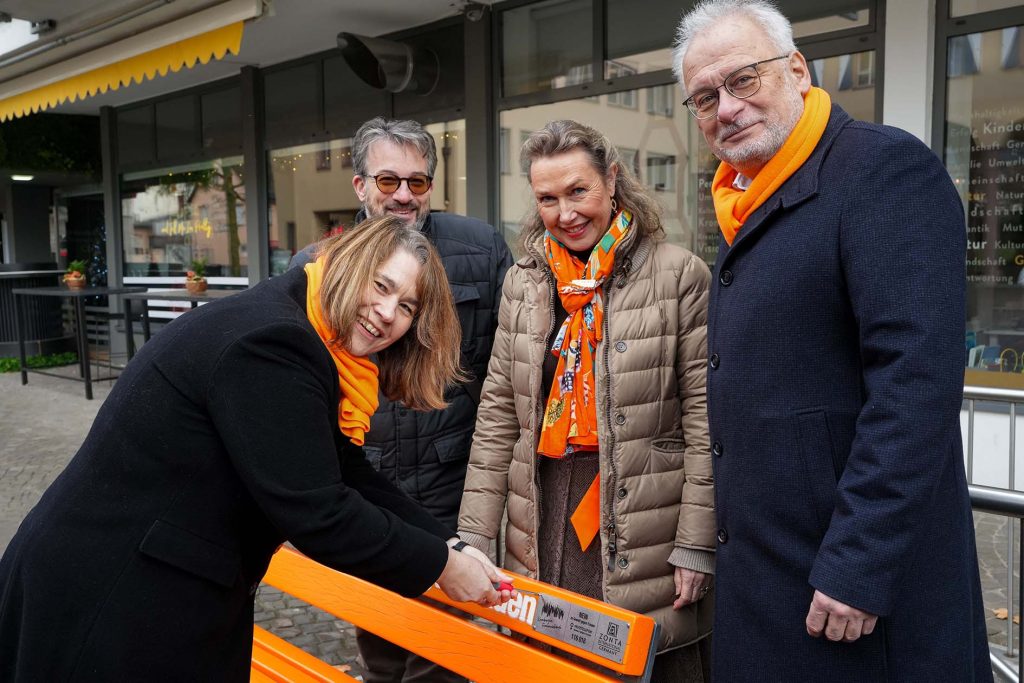 Dr. Ulrike Zymolka befestigte gemeinsam mit Stadtverordnetenvorsteher Andreas Knoche, Zonta-Vertreterin Dr. Ulrike Bilstein und Bürgermeister Christoph König (v.li.) eine Plakette an der orangefarbenen Bank, auf der die Notfallnummer zu finden ist. - Foto: Stadt Kronberg